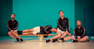 Picture from the performance: four men in sport suits - two of them are sitting at the ends of a gymnastic bench, one man is lying between them and one man is sitting on a floor