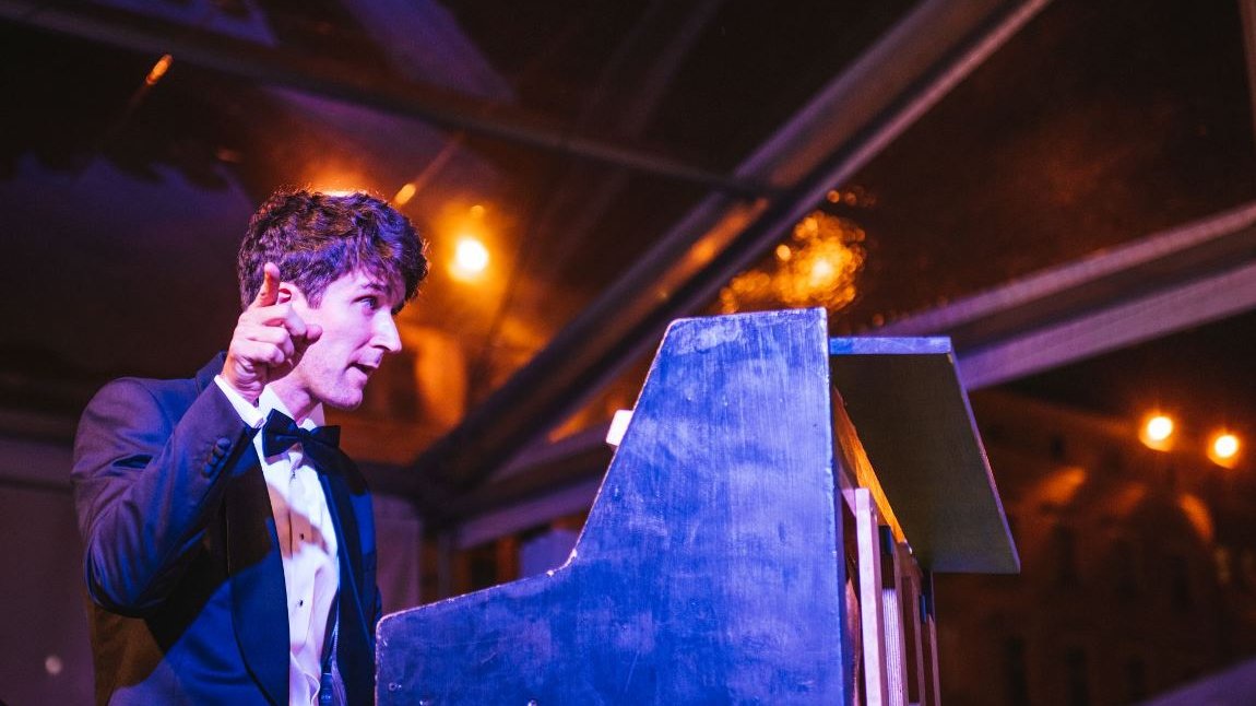 Photo of a man in a suit sitting at the piano and pointing at something with the finger of his right hand - grafika artykułu