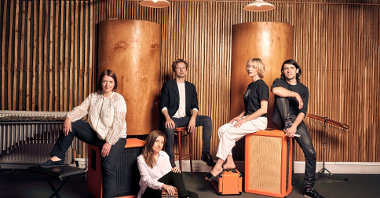 Picture of a band: three women and two men - four of them sitting ond chairs and a box that look like a loudspeaker, one woman is sitting on a floor. The brown-orange wall with two columns in a background.
