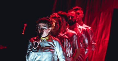 Picture from the performance: 5 people standing in a row. The first one is wearing silver clothes and is holding a microphone to which she is speaking. The other people lighted by red light.
