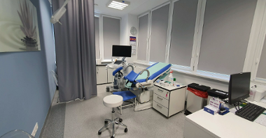 Doctor's office in gray, white and blue colors. On the right the doctor's desk with an open laptop, behind it a cabinet and a chair for gynecological examinations. Behind it a stand with an ultrasound machine.