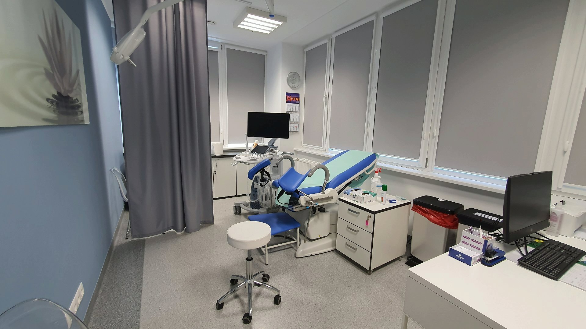 Doctor's office in gray, white and blue colors. On the right the doctor's desk with an open laptop, behind it a cabinet and a chair for gynecological examinations. Behind it a stand with an ultrasound machine.