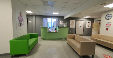 Two lilac couches are set up in the waiting room to the right. Opposite sits the registration desk, parts lowered for people who are short or in wheelchairs. Behind the registration desk is a window. On the left a green couch. Informational posters on the walls.
