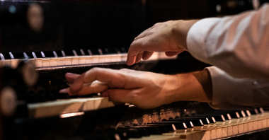 The photo shows a musician's hands on a pipe organ keyboard