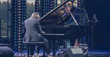 Photo of Leszek Możdżer sitting back to the camera and playing piano on an open-air stage