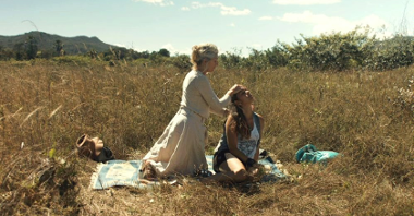 Photo from the movie: two women sitting on a blanket on a field covered with grass and bushes.