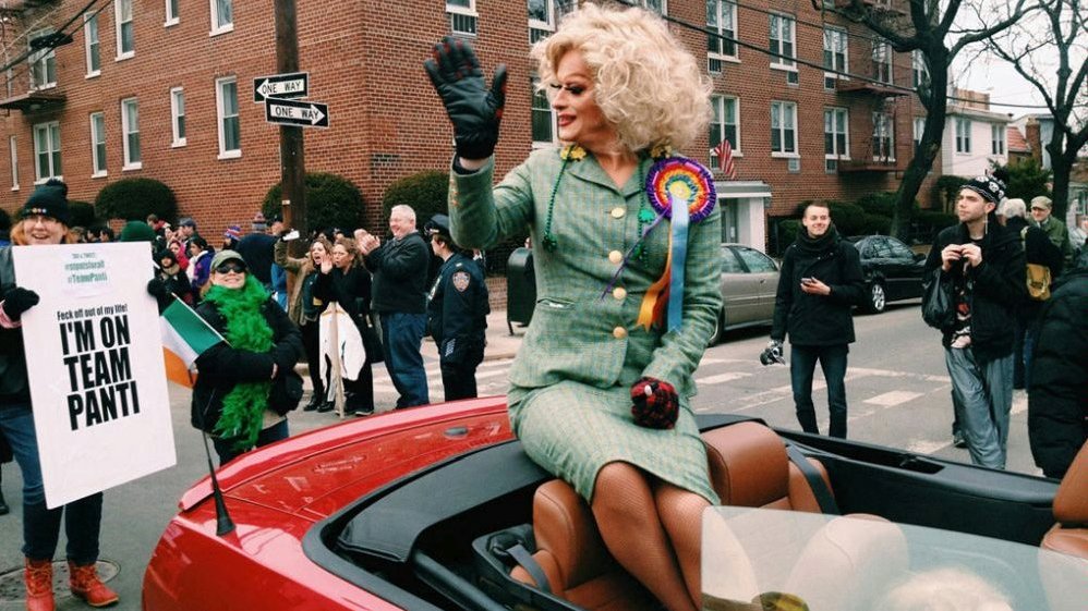 Photo from the movie: a woman sitting on a cabrio's chair. In the background a building and some people gathered on the street