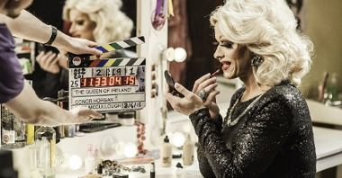 A blond woman in black dress who is sitting by the dressing table and putting her lipstick on. On the left - a man holding a clapperboard.