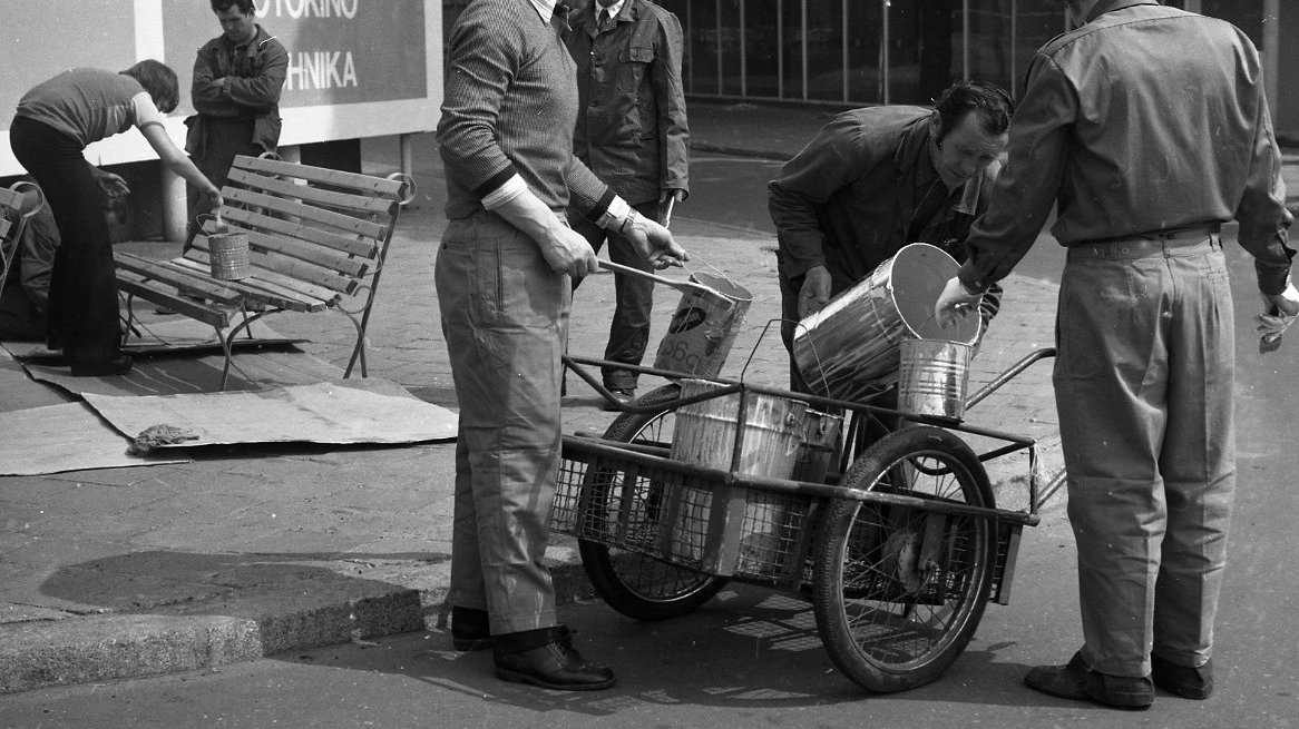 Black and white picture of a few men who are renovating a bench - one of them is paining it, one is standing by a bench, three other men are pouring the paint from big to small can.