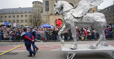 Photo from previous St. Martin parade: picture of St. Martin street with performers (two men pulling a big statue of silver Pegasus on a platform) and people watching the show. The Castle Culture Centre in the background.