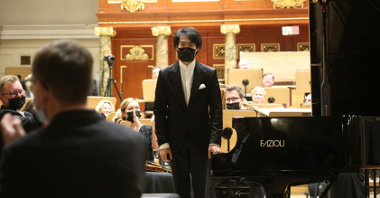Picture from the concert: Bruce Liu standing next to the grand piano. THe members of the orchestra in the background.