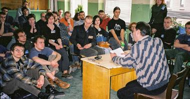 A group of people sitting and listening to the lecture in a classroom. In the foreground a man sitting at the desk, holding papers in his hands.