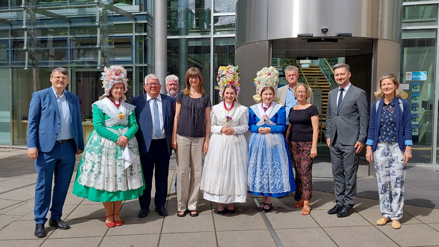 Photo shows people standing in front of office building. Some people wear formal or semi-formal clothing, while few women wear traditional Bambers clothes. - grafika artykułu