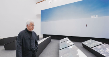 Photo of Kenya Hara standing by his works displayed on low expositors. In the background a big picture in blue and white colours with inscription "Muji".
