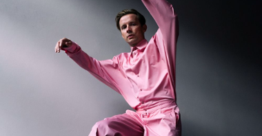 Photo of Bartosz Wąsik - a young man in pink shirt and trousers with his arms and one leg lifted up.