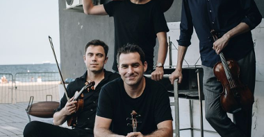 Photo of Atom String Quartet - a four-men band, two of them sitting, another two standing; each of them is holding a musical instrument. A wall of a building and the sea-side as a background.