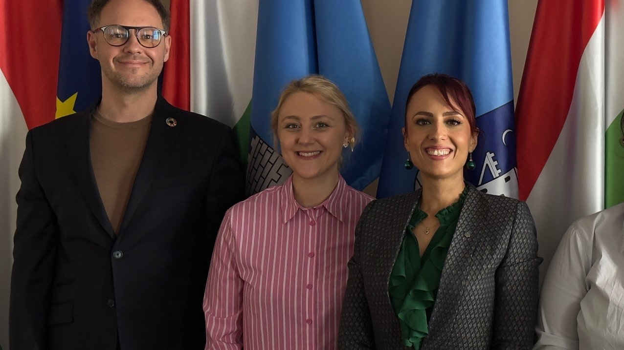 The photo shows three smiling women and a man against a background of several flags