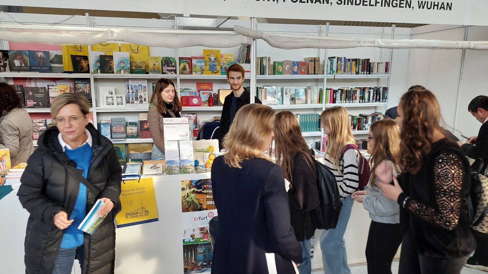 A booth at a cultural or educational fair featuring books and promotional materials. Behind the counter, two staff members (a woman and a man) smile as they engage with visitors. The booth shelves are lined with colorful books and brochures. In the foreground, a group of visitors, mostly women and girls, browse the displayed materials. The setting is well-lit, bustling, and inviting.