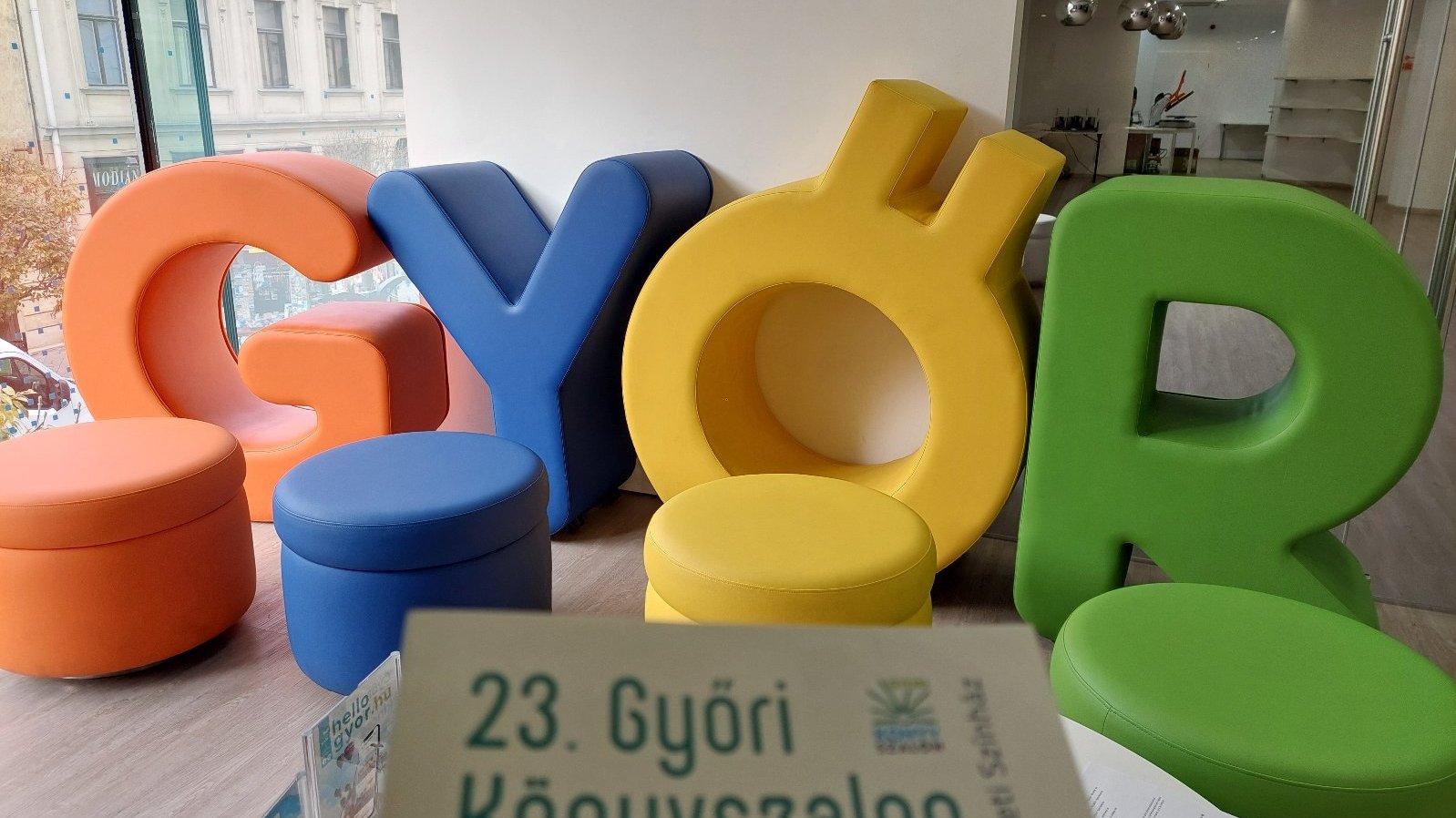 A colorful display from the 23rd Győr Book Fair. In the foreground, a program booklet with the title "23. Győri Könyvszalon" (23rd Győr Book Fair) is partially visible. Behind the letters, there is a glass wall offering a view of a historic building outside. The indoor space is modern and well-lit, with clean, minimalist decor.