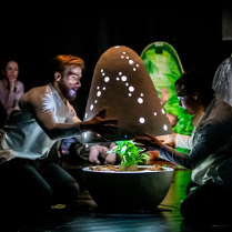 Dwoje ludzi otwiera świecącą kapsułę w której znajduje się roślinka. W tle można zaobserwować inne osoby które patrzą na całość z zaciekawieniem.