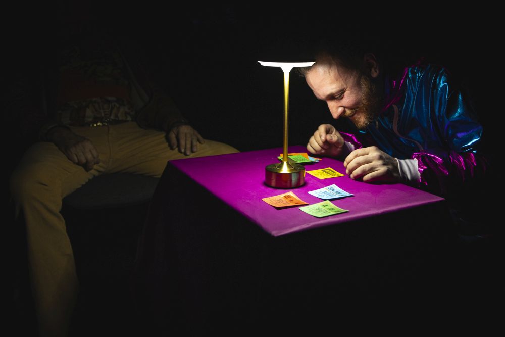 Bohater w siwej, nastroszonej peruce i kolorowym ubraniu siedzi na czerwonej kanapie nonszalancko.