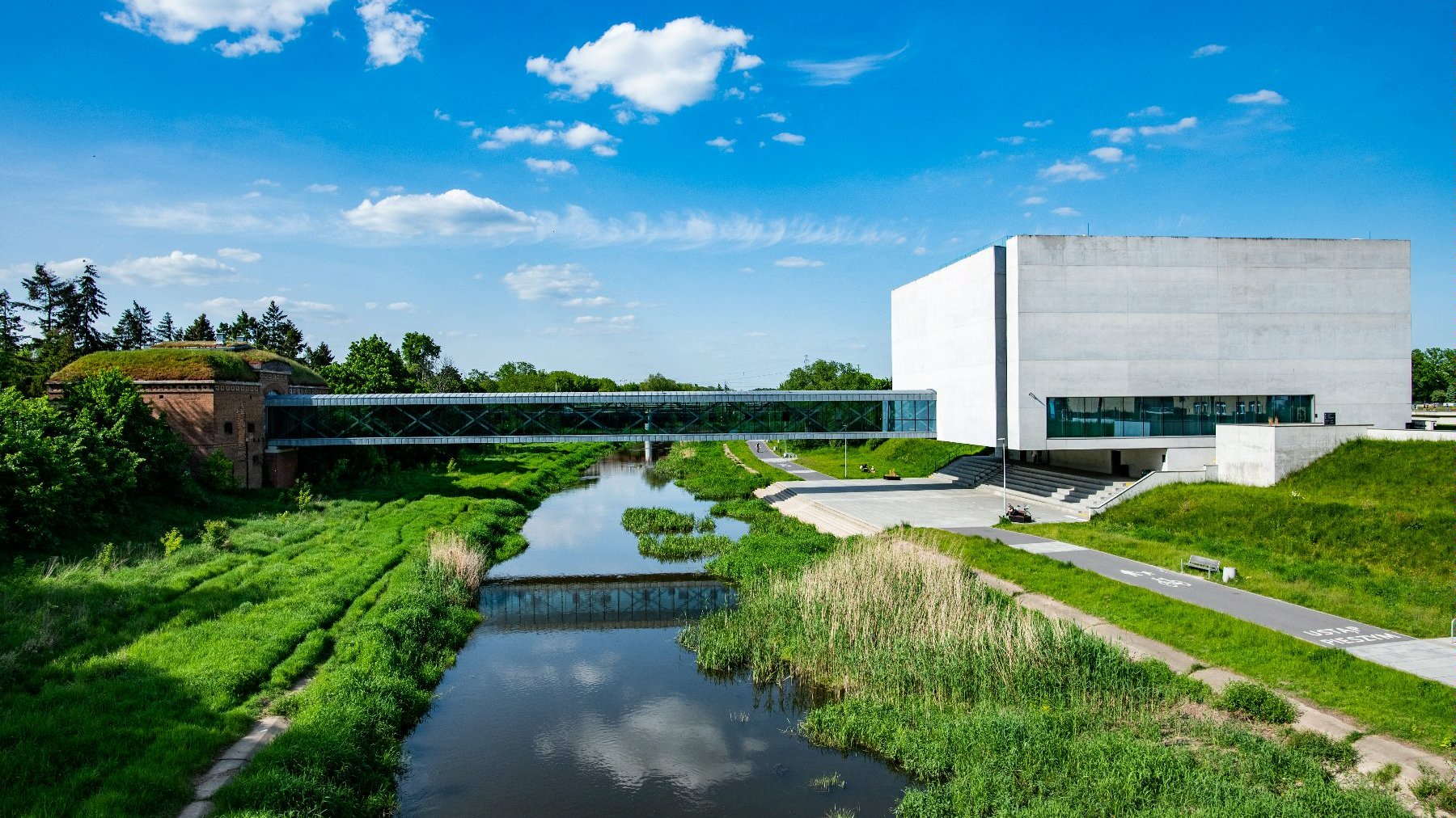 Galeria zdjęć przedstawia prostą, betonową bryłę Bramy Poznania położoną nad rzeką.