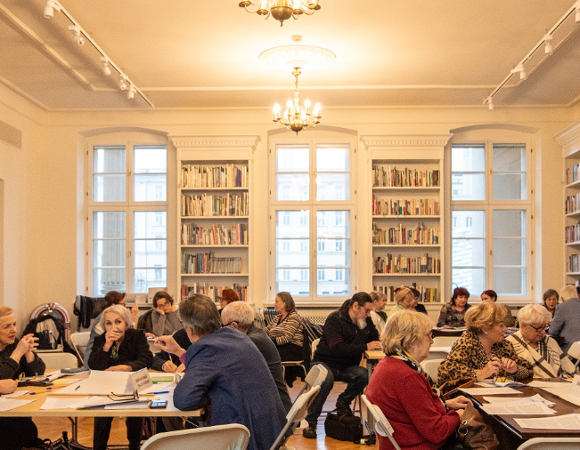 duża jasna sala biblioteki z dużą liczbą stolików, przy których siedzą seniorki i seniorzy. Trwają warsztaty i praca w grupach.