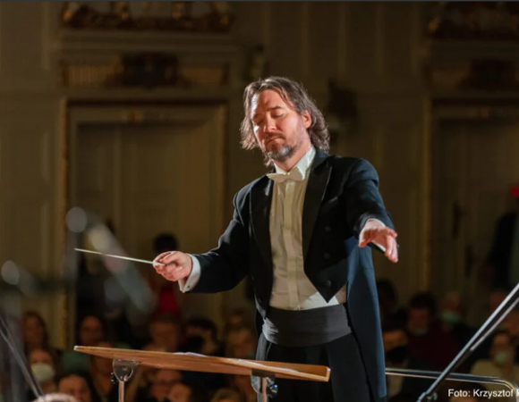 Dyrygent w eleganckim fraku prowadzący orkiestrę podczas koncertu w zabytkowej sali. W tle widoczna publiczność i ciepłe, sceniczne oświetlenie.