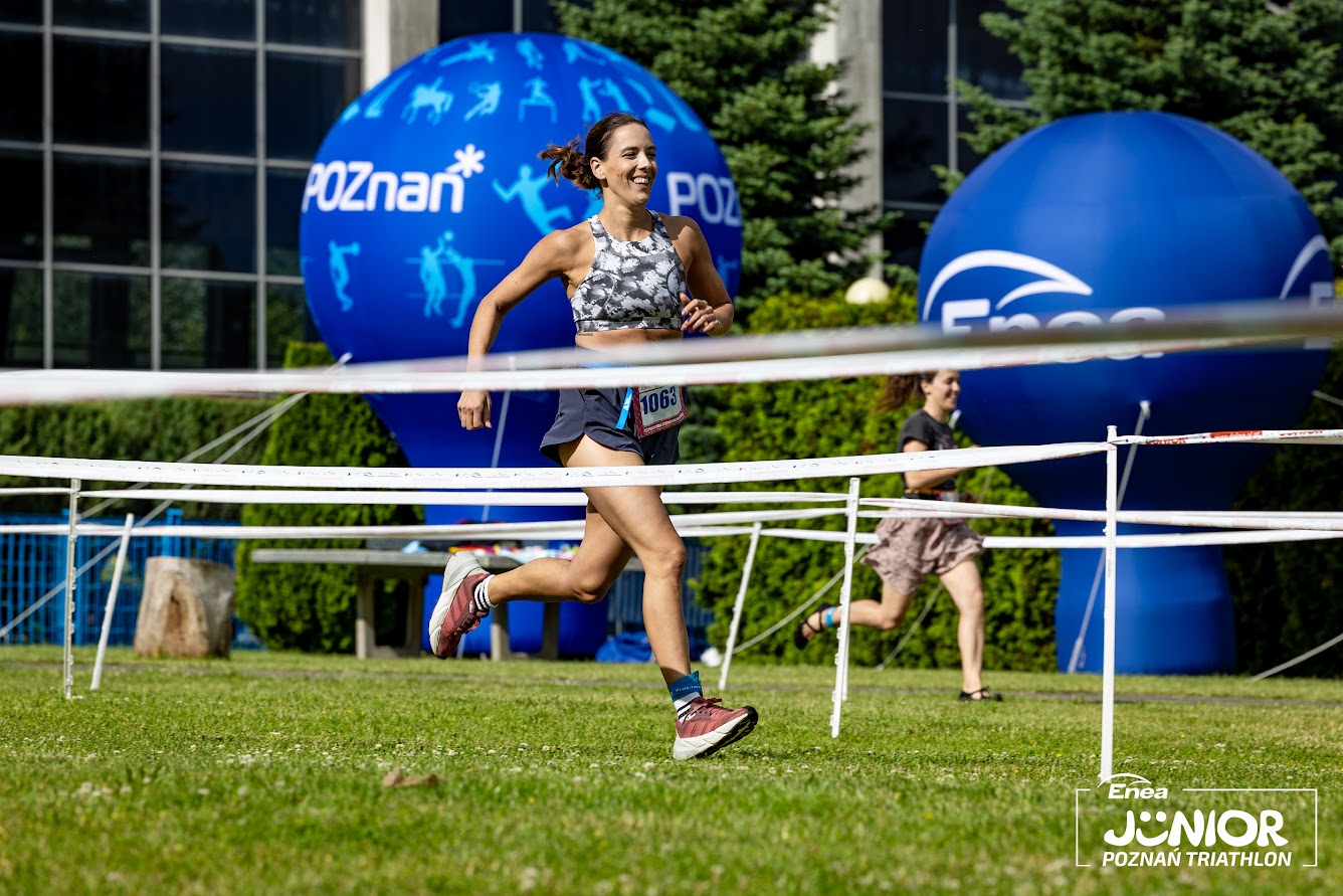 Enea Junior Poznań Triathlon 2024, fot. Jarosław Koperski Enea Junior Poznań Triathlon 2024, fot. Jarosław Koperski - uśmiechnięta kobieta biegnie po trawie na tle niebieskich balonów Poznania i Enei. W oddali widać także inną biegnącą kobietę