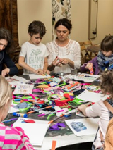 Opiekunowie z dziećmi na zajęciach przy stole pełnym kartek i kolorowych papierów.