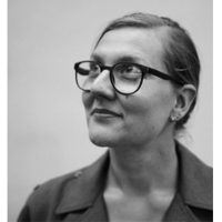 Na zdjęciu jest Marta Madejska, w portretowym ujęciu. Ma związane, gładko zaczesane włosy, okulary w wyrazistych oprawkach i marynarkę. Jej głowa skierowana jest w lewo, patrzy w górę z delikatnym uśmiechem.