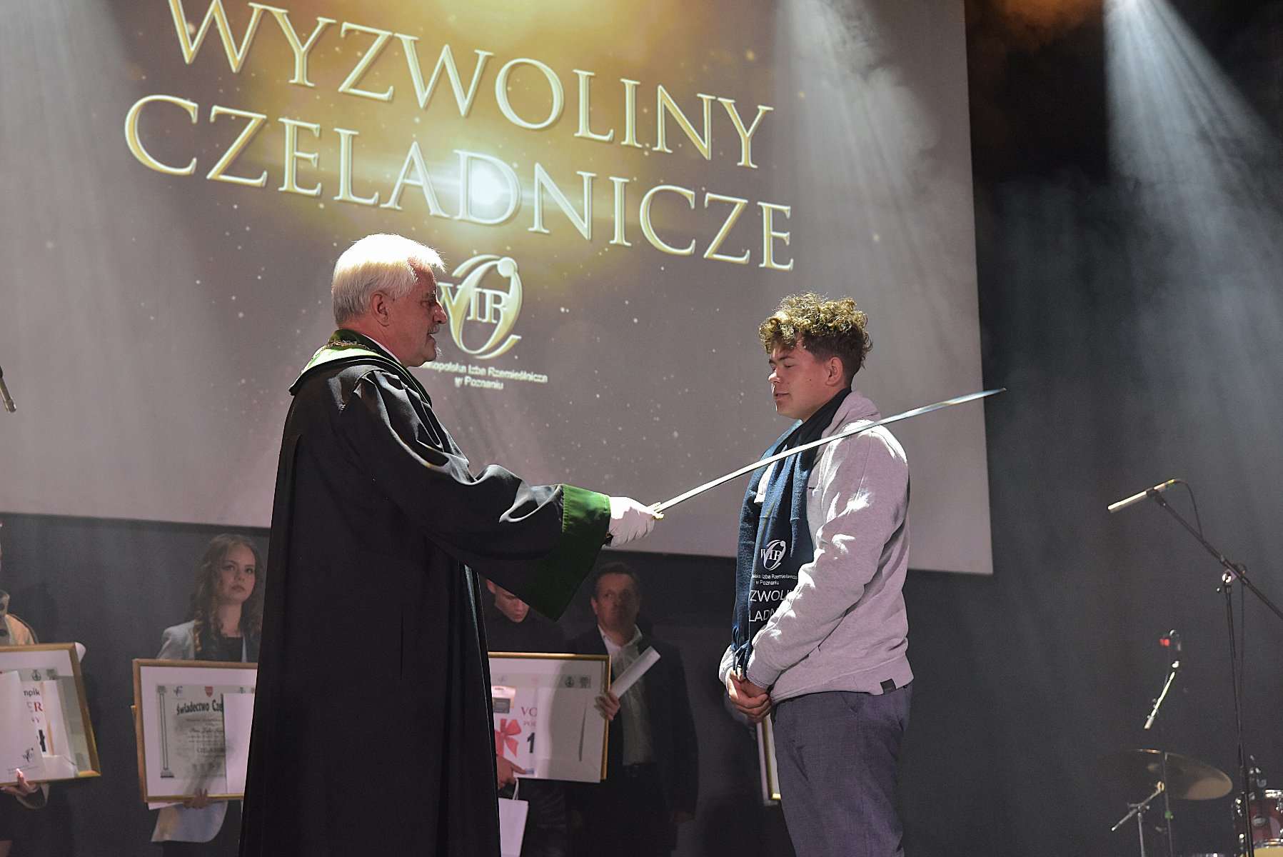 Galeria zdjęć z uroczystości Wyzwolin czeladniczych - Na zdjęciu widać uroczystość wręczenia dyplomów czeladniczych - mężczyzna w todze symbolicznie pasuje mieczem młodego absolwenta stojącego na scenie. W tle widoczny jest napis "Wyzwoliny Czeladnicze