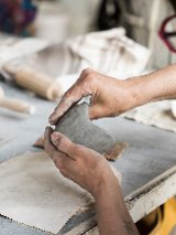 Kobieta modelująca ręcznie gliniany przedmiot na stole warsztatowym, otoczona narzędziami do ceramiki i materiałami do pracy.