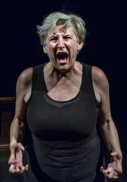 na zdjęciu kobieta w średnim wieku patrzy w obiekty i krzyczy głośno, ręce ma skierowane do przodu, place zagięte