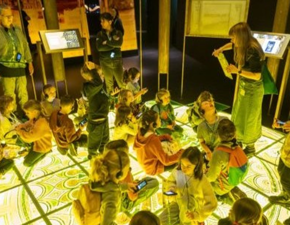 Grupa dzieci podczas zajęć w Bramie Poznania. Dzieci siedza na podświetlonej podłodze, wokół nich stoi kilkoro dorosłych.