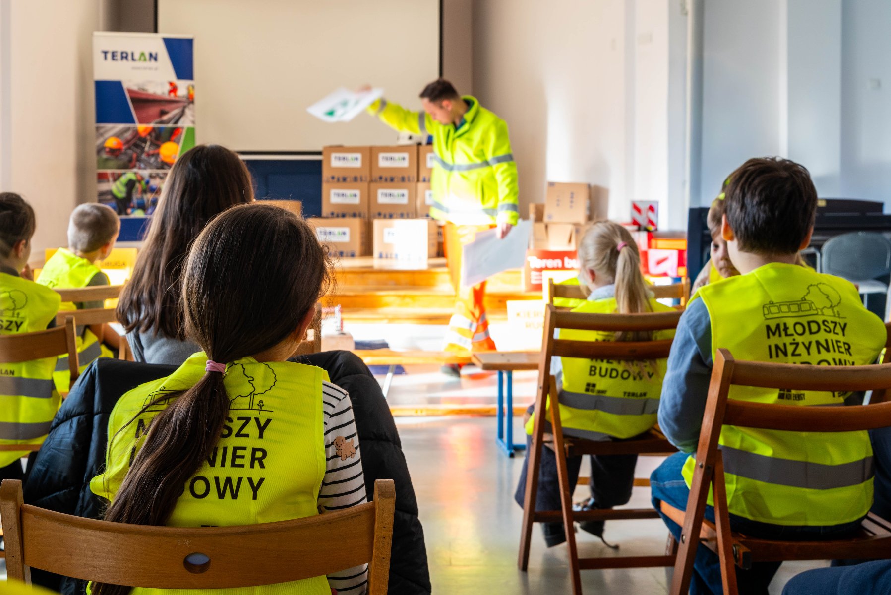 Grupa dzieci w żółtych kamizelkach odblaskowych siedzi na krzesłach i słucha prezentacji prowadzonej przez osobę w jaskrawej odzieży roboczej. Przed prowadzącym stoją ułożone kartony z logo firmy TERLAN, a w tle znajduje się ekran projekcyjny.