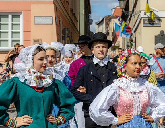 W niedzielę 3 sierpnia ulicami Starego Rynku przejdzie barwny korowód Bambrów Grupa ludzi na Starym Rynku przebranych w tradycyjne bamberskie stroje, m.in. kolorowe sukienki i ozodby z kwiatów lub białe czepki na głowie. Za nimi tłum innych ludzi.