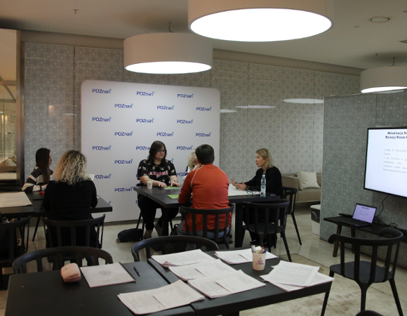grupa ludzi siedzących przy stole biorących udział w warsztatach. Na zdjęciu widoczne są materiały papierowe do wykorzystania podczas warsztatów, ekran z prezentacją multimedialną oraz ścianka z logo Poznania