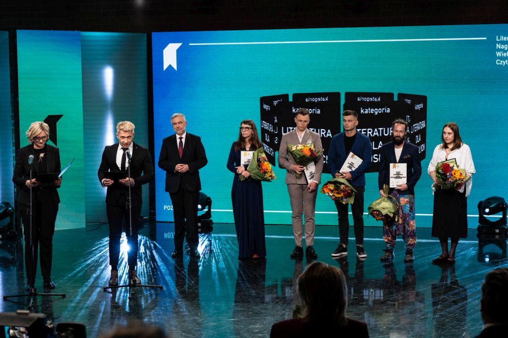 fot. WBPiCAK Grupa ludzi stoi na scenie trzymając dyplomy i bukiety kwiatów. Dwoje prowadzących galę wręczenia nagród przemawia do mikrofonów na statywach.