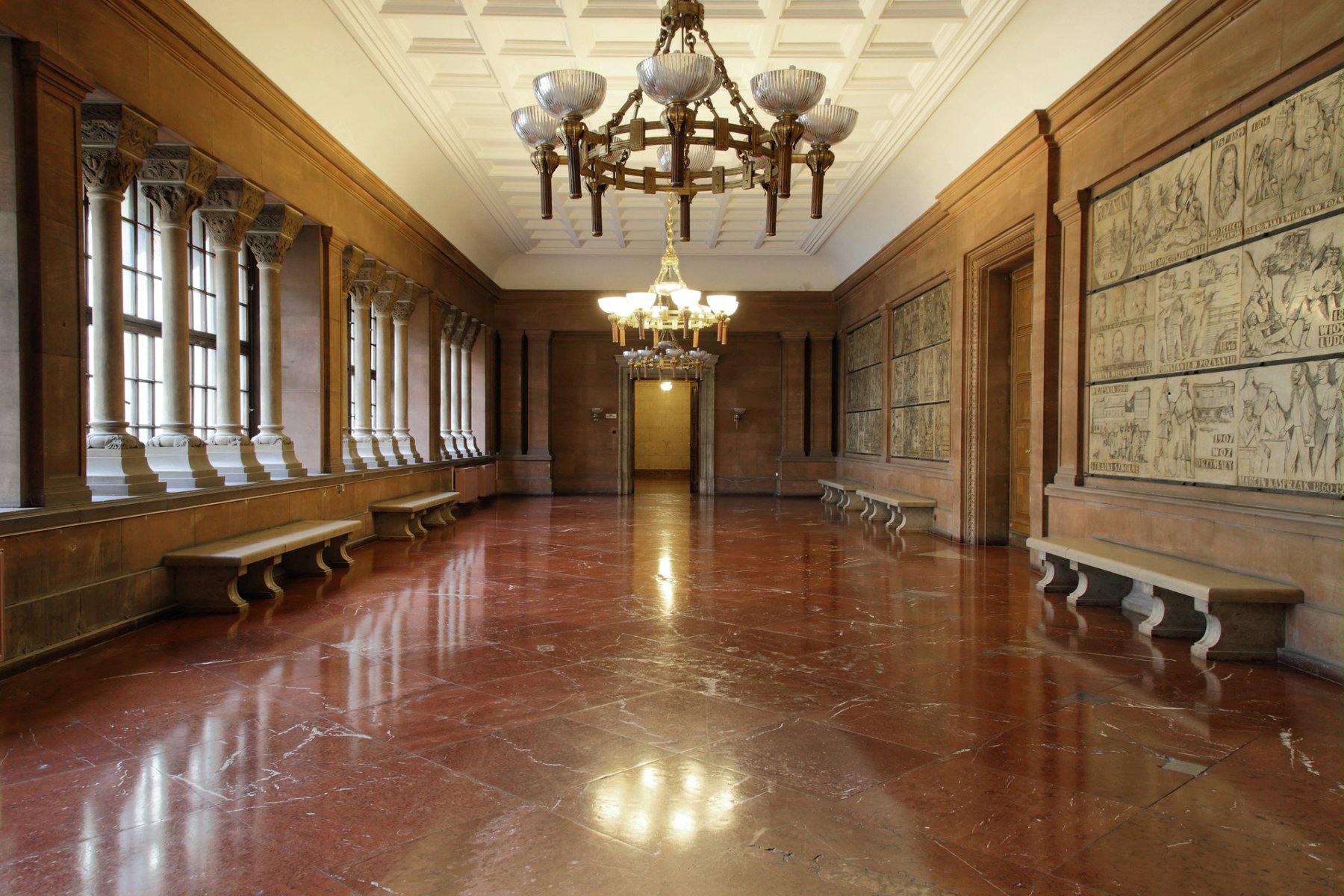 Zdjęcie jednego z zamkowych korytarzy. Kamienna posadzka w ciemnoczerwonym kolorze, okazałe żyrandole zwisające z sufitu. Z lewej strony rząd okien, pod ścianami ławki.