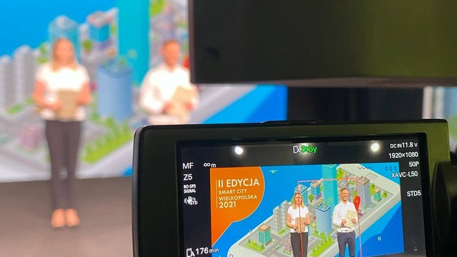 Zdjęcie widoku z kamery, która filmuje prowadzących konferencję. Dwie osoby stojące, w tle kolorowa grafika.
