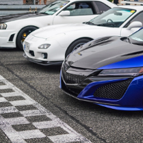 Na linii startowej stoją trzy sportowe samochody. Dwa z nich są białe a jeden czarno-granatowy.