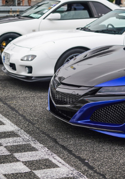 Na linii startowej stoją trzy sportowe samochody. Dwa z nich są białe a jeden czarno-granatowy.