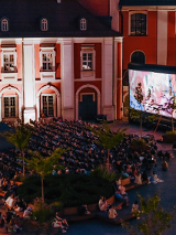 Widok z góry na plac Kolegiacki, na którym stoi duży ekran kinowy i rozłożone leżaki. Na ekranie wyświetla się film. Na leżakach siedzi widownia. Wokół małe drzewka. W tle budynek Kolegiaty.