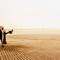 Wejście na plażę z drewanianego molo. Po lewej przy płocie nachylający się mężczyzna do mini fonnanty z wodą pitną.