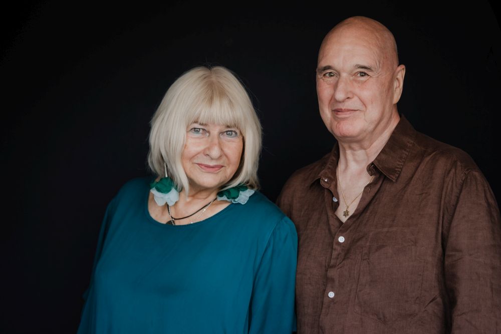 Elżbieta "Bieta" Ficowska i Cezary Harasimowicz, fot. Maciej Zienkiewicz Photography Kobieta i mężczyzna w średnim wieku pozują razem do zdjęcia. Ona ma blond włosy, grzywkę i duże kolczyki w kształcie kwiatów, on jest łysy, w brązowej koszuli rozpiętej pod szyją.