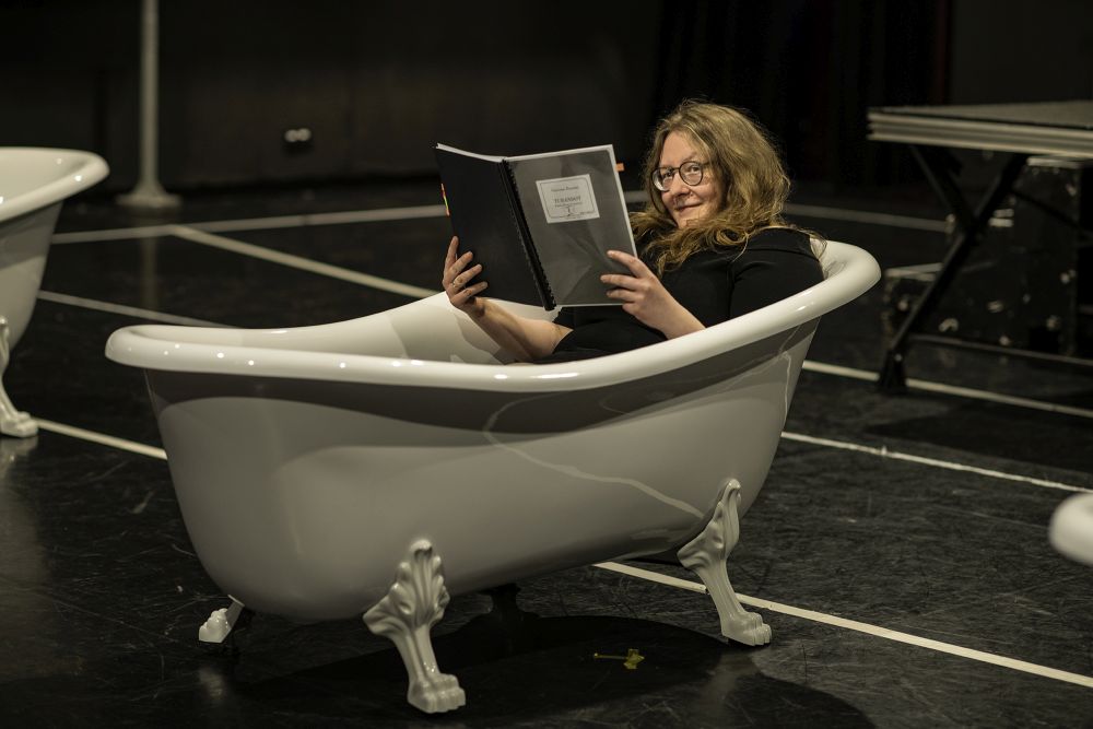 Kobieta leży w zdobionej, staroświeckiej wannie, która stoi na scenie jako rekwizyt. Uśmiecha się, czyta duży zeszyt.