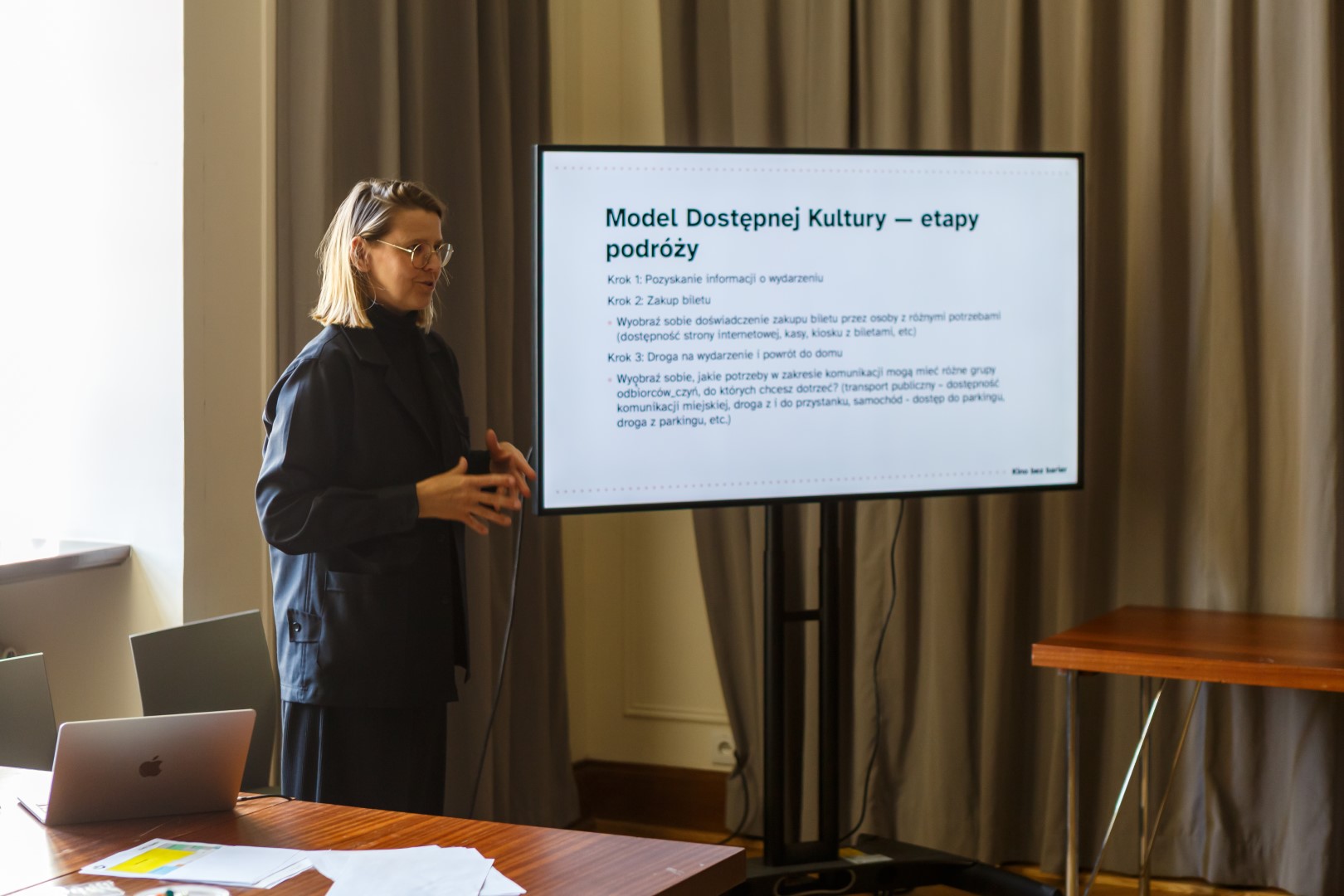 Kobieta prowadzi prezentację. Obok niej widoczny ekran ze slajdem prezentacji o dostępności kultury.