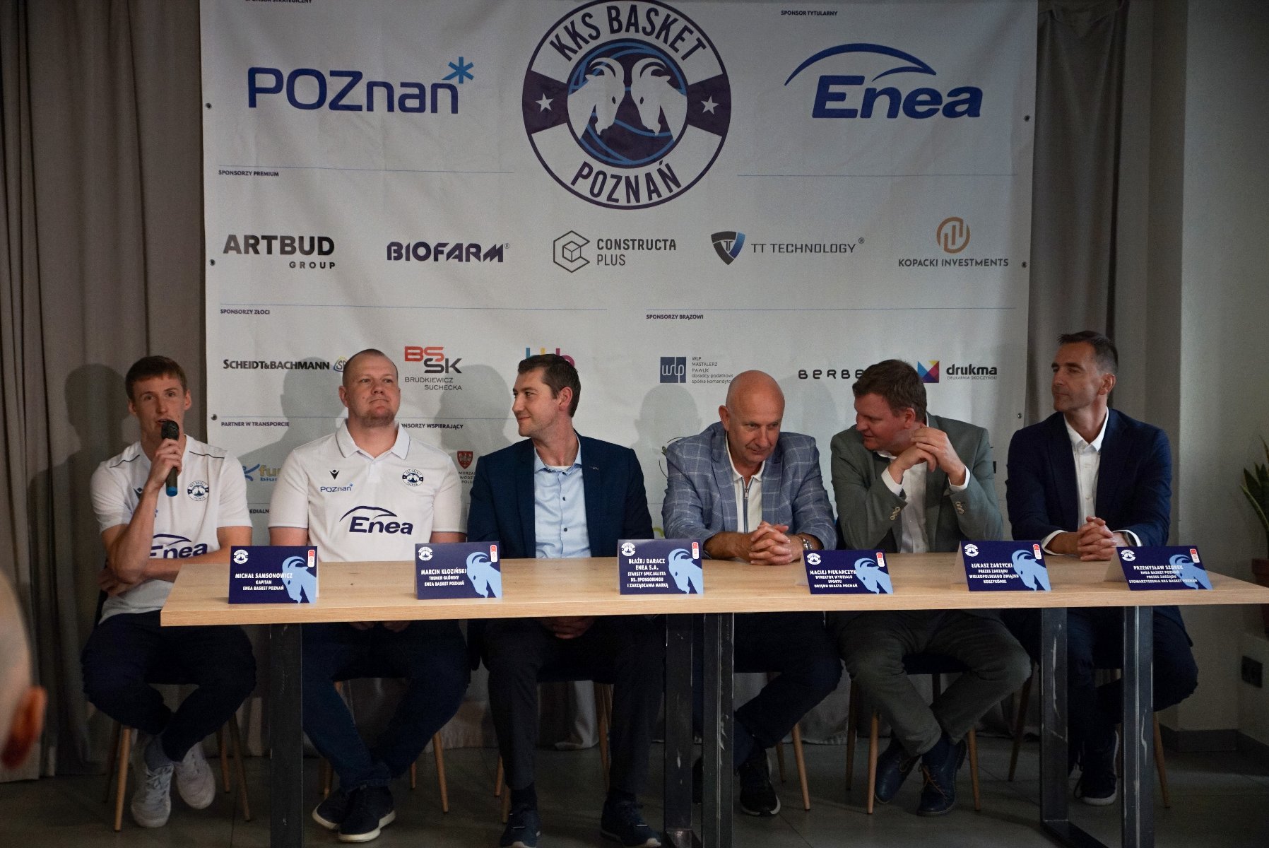 Konferencja prasowa przed sezonem Enea Basket Poznań, fot. Beata Brociek Konferencja prasowa przed sezonem Enea Basket Poznań, fot. Beata Brociek - za stołem siedzi 6 mężczyzn na tle klubowego baneru. Mężczyzna z lewej strony trzyma mikrofon