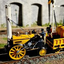 To jest miniaturowy model parowozu Rocket, historycznego pociągu, który zrewolucjonizował transport. Został ustawiony na makiecie torów kolejowych, kompletny z miniaturowym wagonem węglowym i figurkami ludzi. Model jest bardzo szczegółowy, z dokładnymi reprezentacjami projektu lokomotywy, w tym kotła, kół i nawet kominu. Model jest również pomalowany w jaskrawych kolorach, co dodaje realizmu. Tło zdjęcia wydaje się być miniaturowym modelem kamiennego muru, z arkadami w tle, dodającym do poczucia skali.